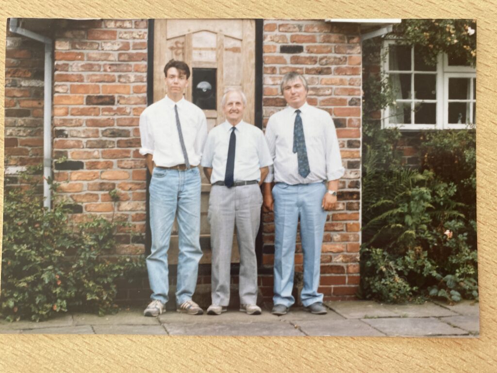 Three men standing outside a nursery with plants and brick building.